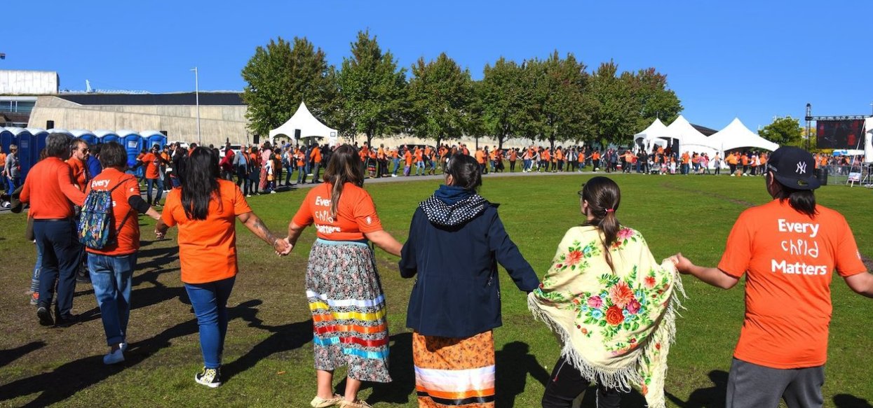 People hold hands in a large circle as drummers perform on stage at an event on National Day for Truth and Reconciliation Day.